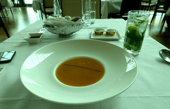 A first-class dining table set with a bowl of orange soup, a mojito-style drink, and a bread basket.