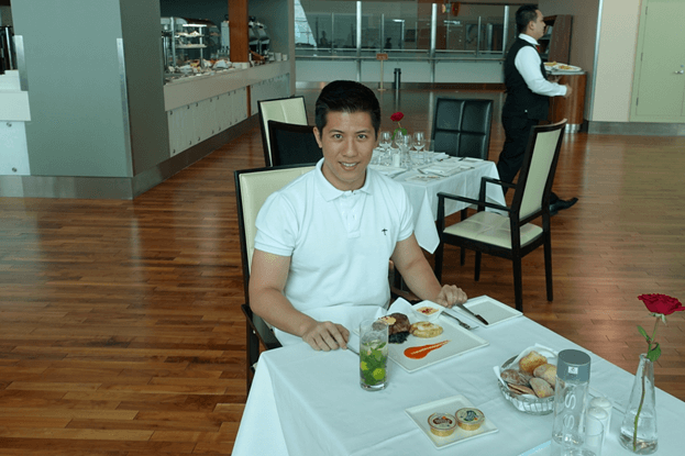 A man smiles while dining at a table in a bright First Class lounge.