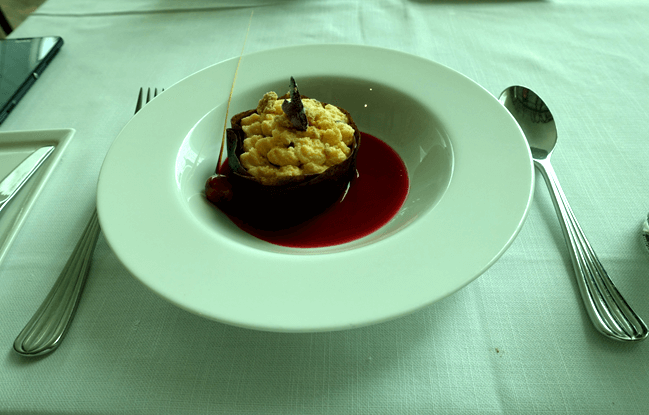 A gourmet dessert in a white bowl featuring a chocolate cup with crumble, red sauce, and a spoon on a white tablecloth.