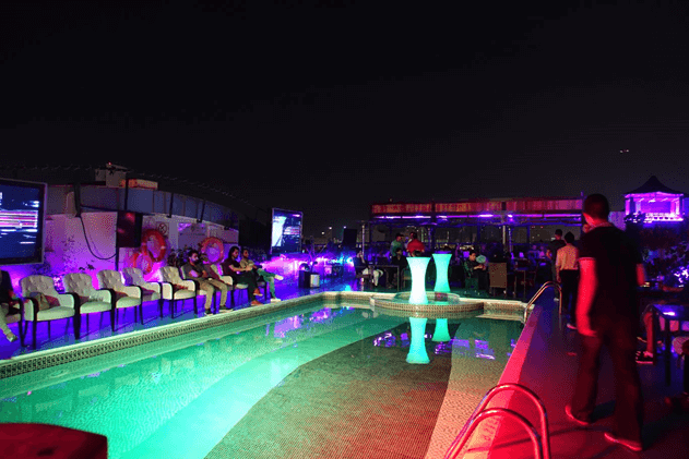 A vibrant rooftop bar at night with a swimming pool, people lounging and watching large screens under colorful lights.