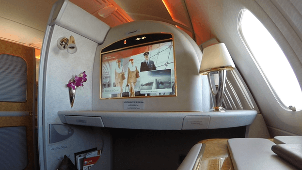 An Emirates A380 First Class suite interior with a large entertainment screen, lamp, and flowers.