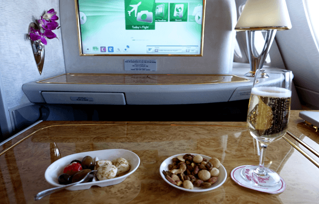 A glass of champagne and snacks on a wood table in an Emirates First Class cabin, with an entertainment screen and orchid vase visible.