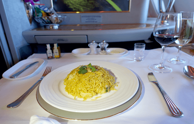 A plate of yellow rice with wine and water is served on a set table in an Emirates First Class cabin.