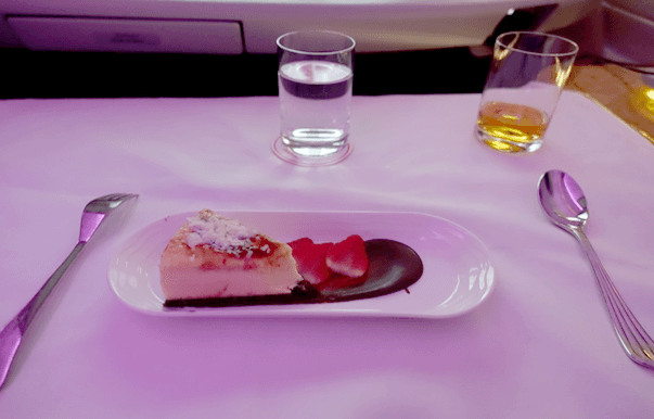 A slice of cheesecake with strawberries, chocolate sauce, drinks, and cutlery on an Emirates First Class table with purple ambient lighting.