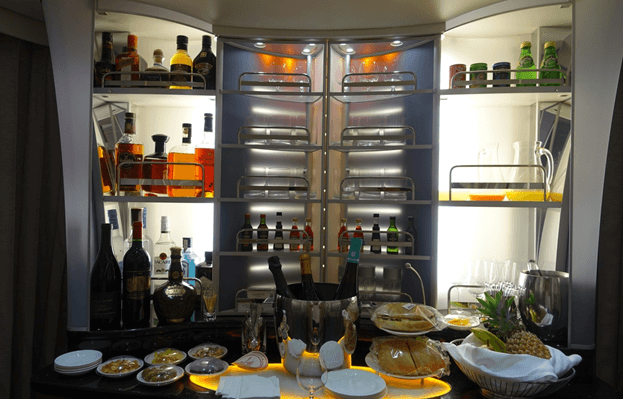 An illuminated onboard bar in Emirates First Class, stocked with various alcoholic drinks, champagne, snacks, and fresh fruit.