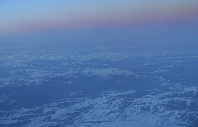 Aerial view of snow-capped mountains and clouds under a hazy blue and purple sky.