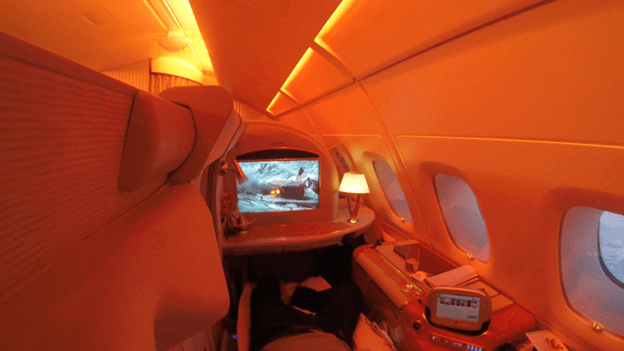 Emirates A380 First Class private suite with a passenger relaxing and watching the entertainment screen in warm orange light.