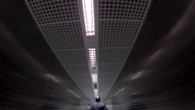 A person walks down a long, reflective airport corridor with a gridded ceiling and fluorescent lights.