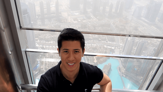 Man takes a selfie from a high observation deck overlooking the Dubai cityscape and a turquoise lake.