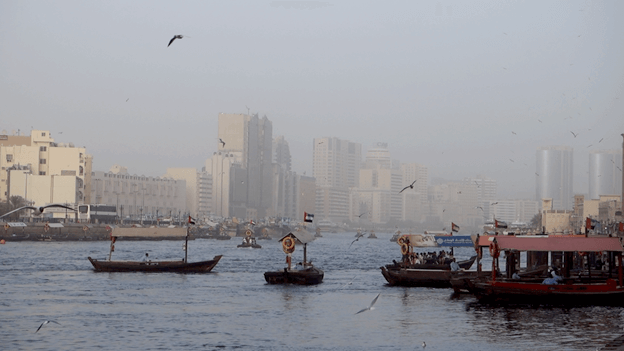 Traditional abra boats on Dubai Creek with a hazy city skyline and flying birds.