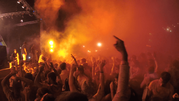 An energetic crowd with hands raised at a concert, illuminated by bright orange lights and smoke from the stage.