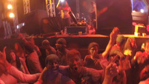 An excited crowd, many with colorful powder on their faces, cheers at a concert or festival.