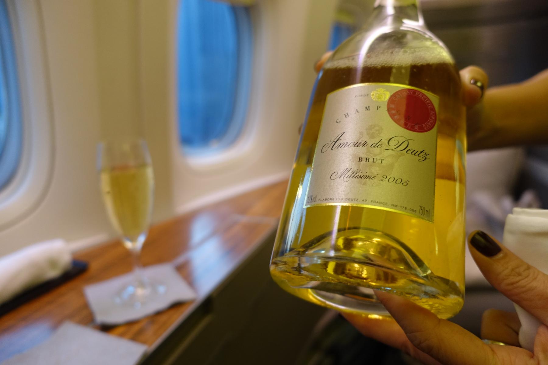 A hand holds a bottle of Amour de Deutz Millésime 2005 champagne in a Cathay Pacific First Class cabin.