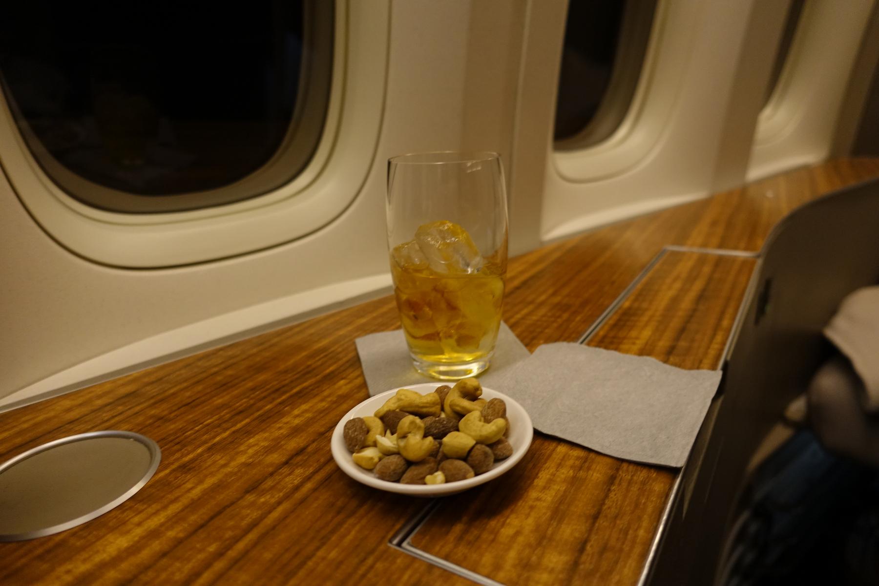 An iced drink and a bowl of mixed nuts are served on a polished wooden table in a first class airplane cabin.