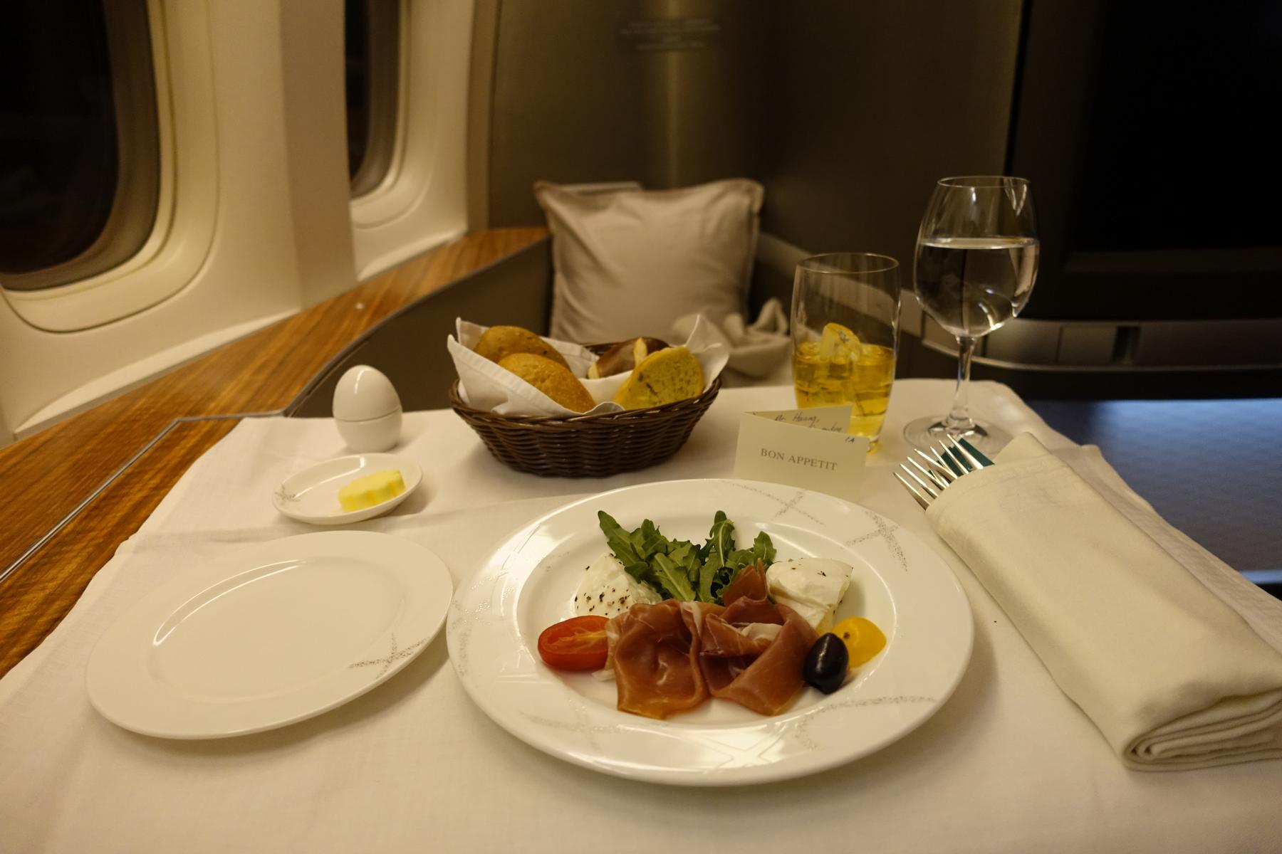 A gourmet meal, including prosciutto and cheese, served on a white tablecloth in an airplane's first class cabin.