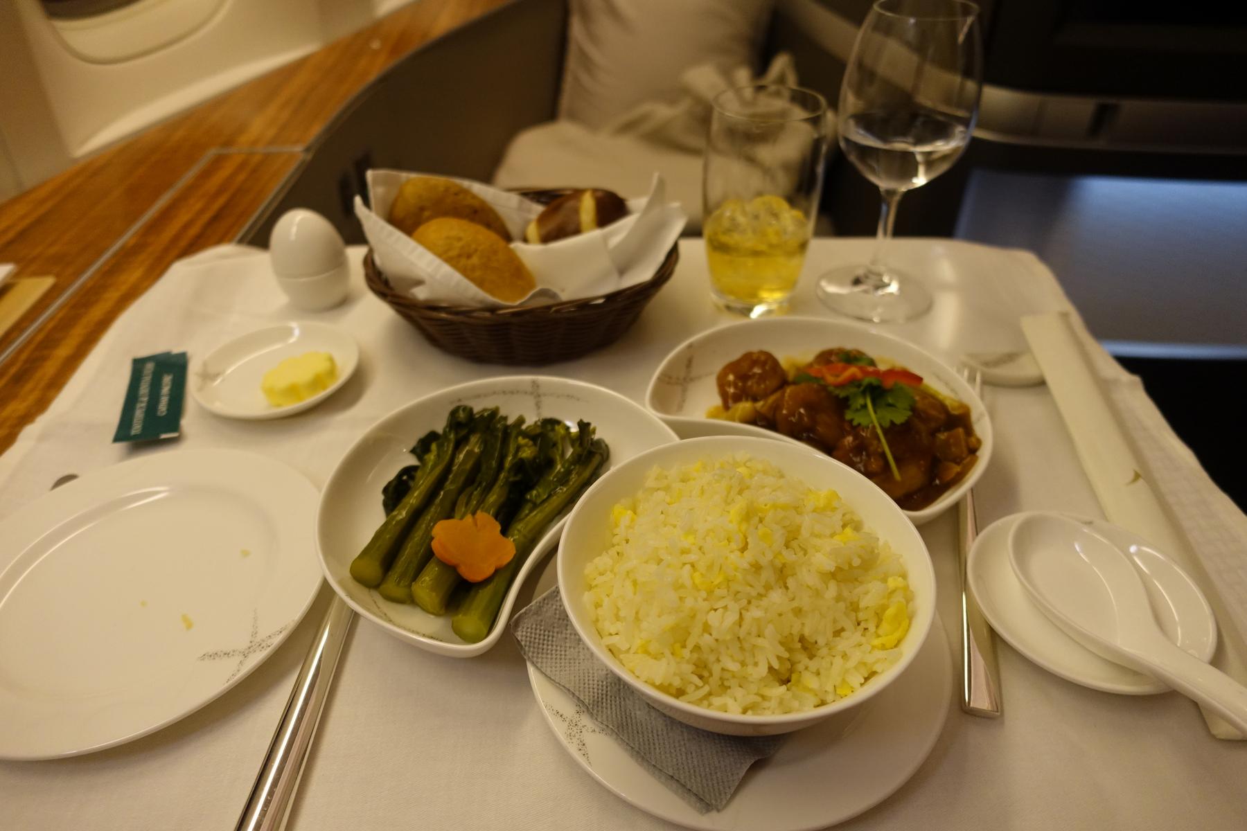 A multi-course First Class meal is elegantly presented on a white tablecloth, featuring egg fried rice, braised meat, green vegetables, and bread.