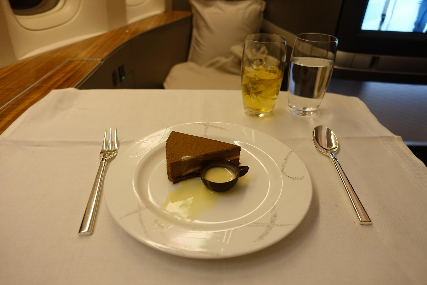 A slice of chocolate dessert with sauce, silverware, and two drinks served on a white tablecloth in a first-class airplane cabin.