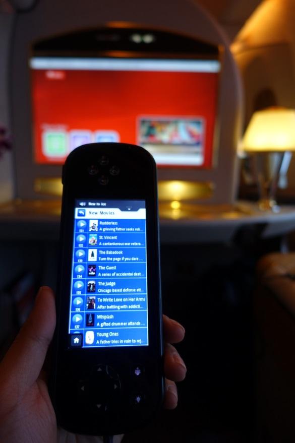 A hand holds a remote control displaying a list of new movies, with an Emirates First Class entertainment screen visible in the background.