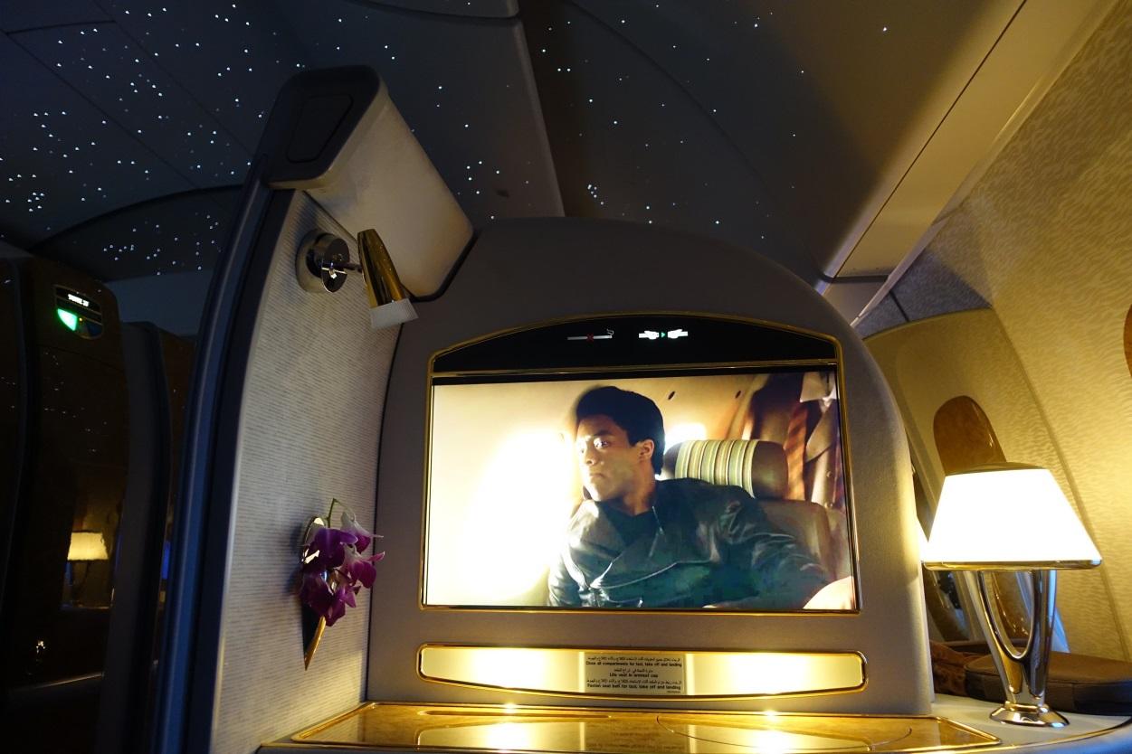 Emirates First Class suite interior with a movie playing on a large screen, a desk lamp, purple orchids, and a starry night ceiling.