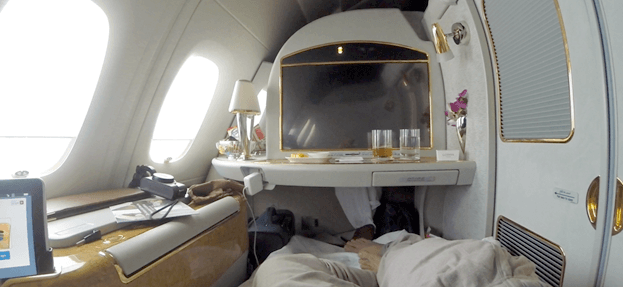 A luxurious Emirates First Class suite on an A380 with a large screen and a passenger relaxing.