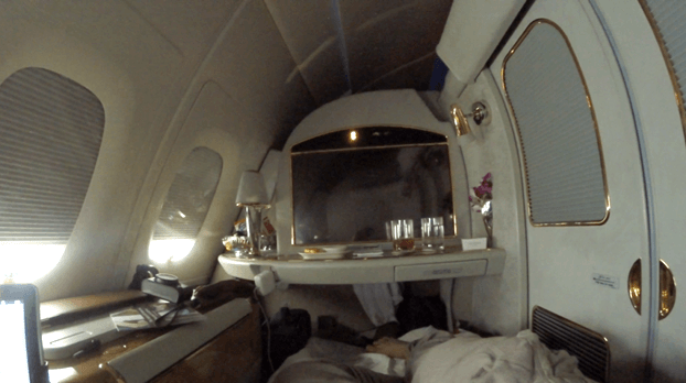 A passenger relaxes in a luxurious Emirates A380 First Class suite, featuring a large screen and drinks.