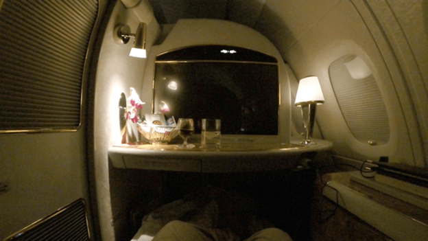 A dimly lit Emirates A380 First Class suite showing a personal screen, lamp, and drinks on a table.