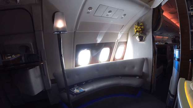 Emirates A380 First Class onboard lounge with curved sofa, floor lamp, and three windows.