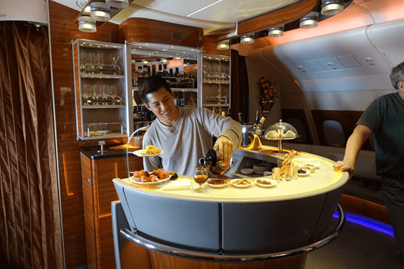 A man smiles while pouring a drink at the fully stocked, luxurious onboard bar of an Emirates A380 First Class flight.