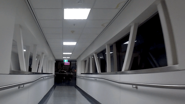 A brightly lit airport jet bridge with white walls, a handrail, and windows looking out to darkness.