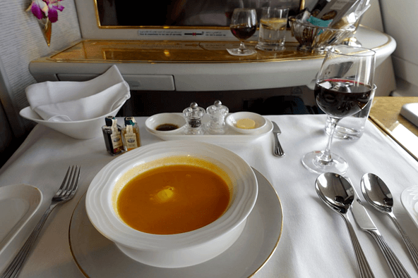 An elegantly plated meal of orange soup, red wine, and condiments is served on a white tablecloth in an Emirates First Class cabin.