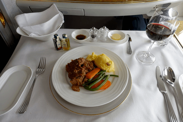 An elegant Emirates First Class meal featuring meat, mashed potatoes, vegetables, and red wine served on a white table.