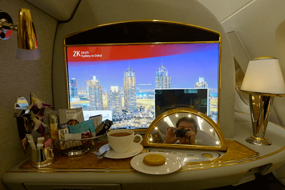 An Emirates A380 First Class suite showing a large screen with a city view and flight info, along with tea, snacks, and a reflection of the photographer.