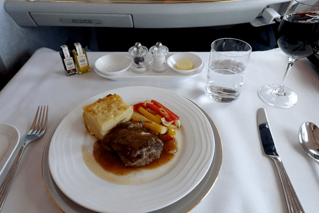 An Emirates First Class meal of meat, potato gratin, vegetables, and wine served on a white tablecloth.