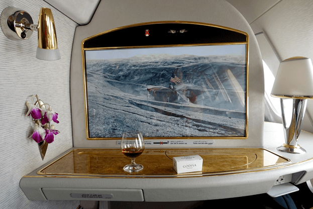 An Emirates First Class suite features a large screen, a glass of amber liquid, Godiva chocolates, and fresh orchids.