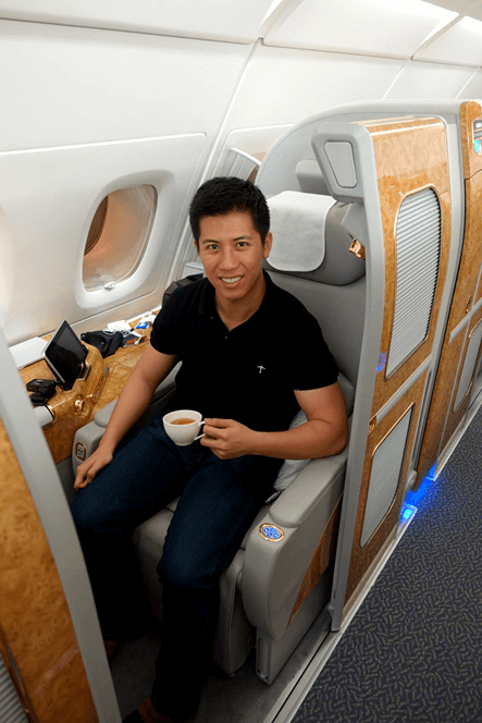 A smiling man sits in an Emirates A380 First Class suite, holding a cup.
