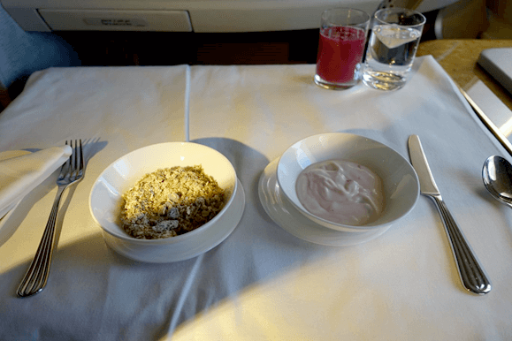 A breakfast meal of granola, yogurt, and drinks served on a white tablecloth in Emirates First Class.