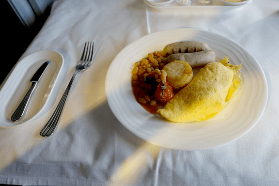 An Emirates First Class breakfast meal with an omelette, sausages, baked beans, and roasted tomato on a white plate.