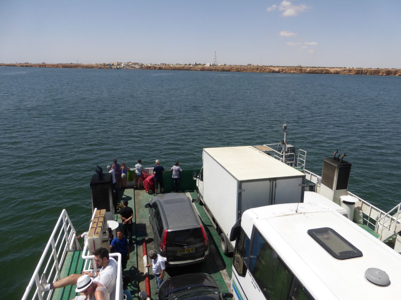 View from a ferry carrying passengers and vehicles, looking across the water towards a distant town on a rocky coastline.