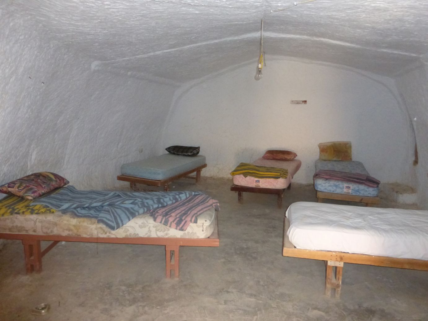 A rustic, white cave-like room features several simple beds and a single hanging lightbulb.
