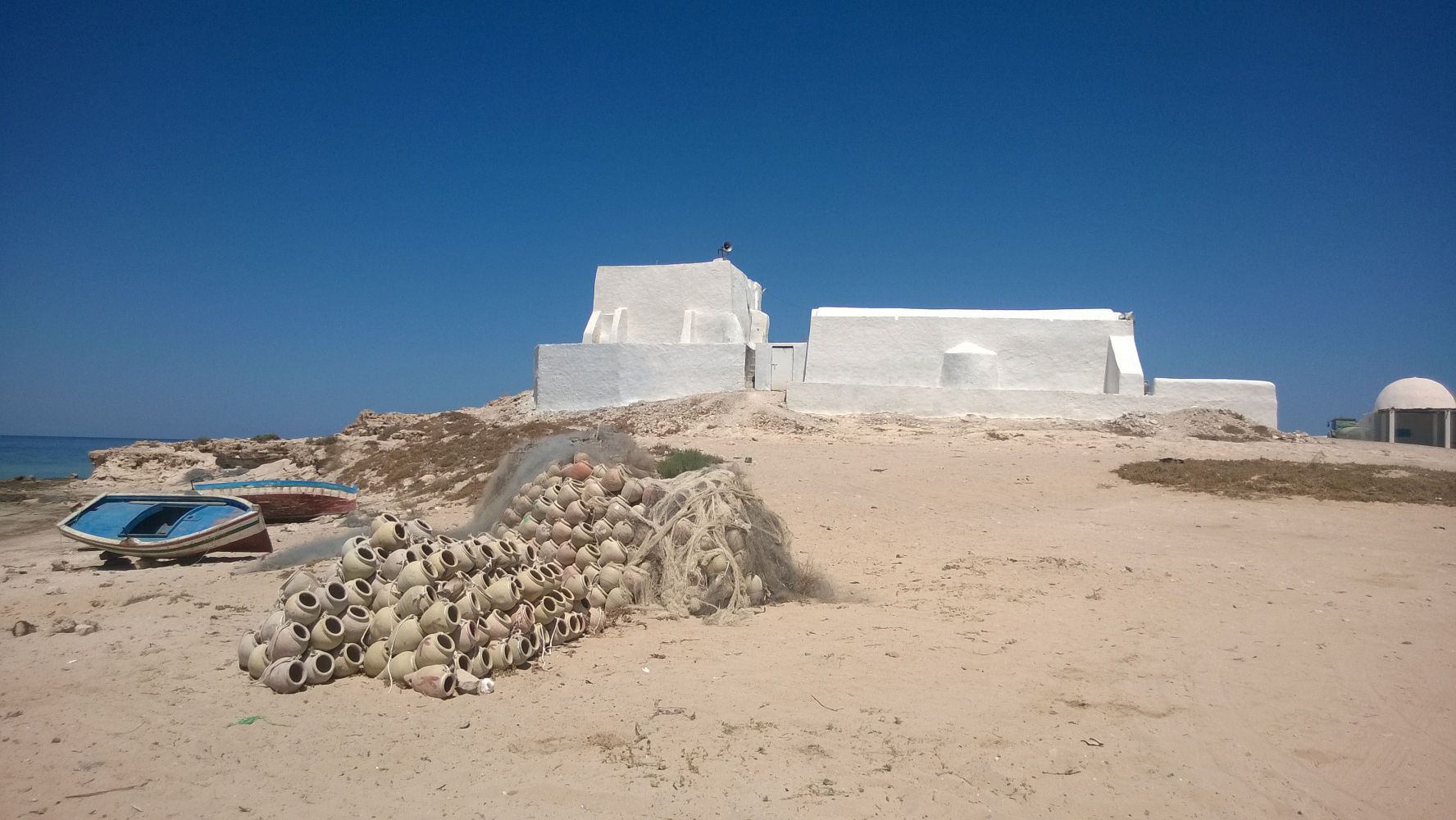 White, geometric buildings from a Star Wars set rise above fishing boats and stacked terracotta pots on a sandy shore.