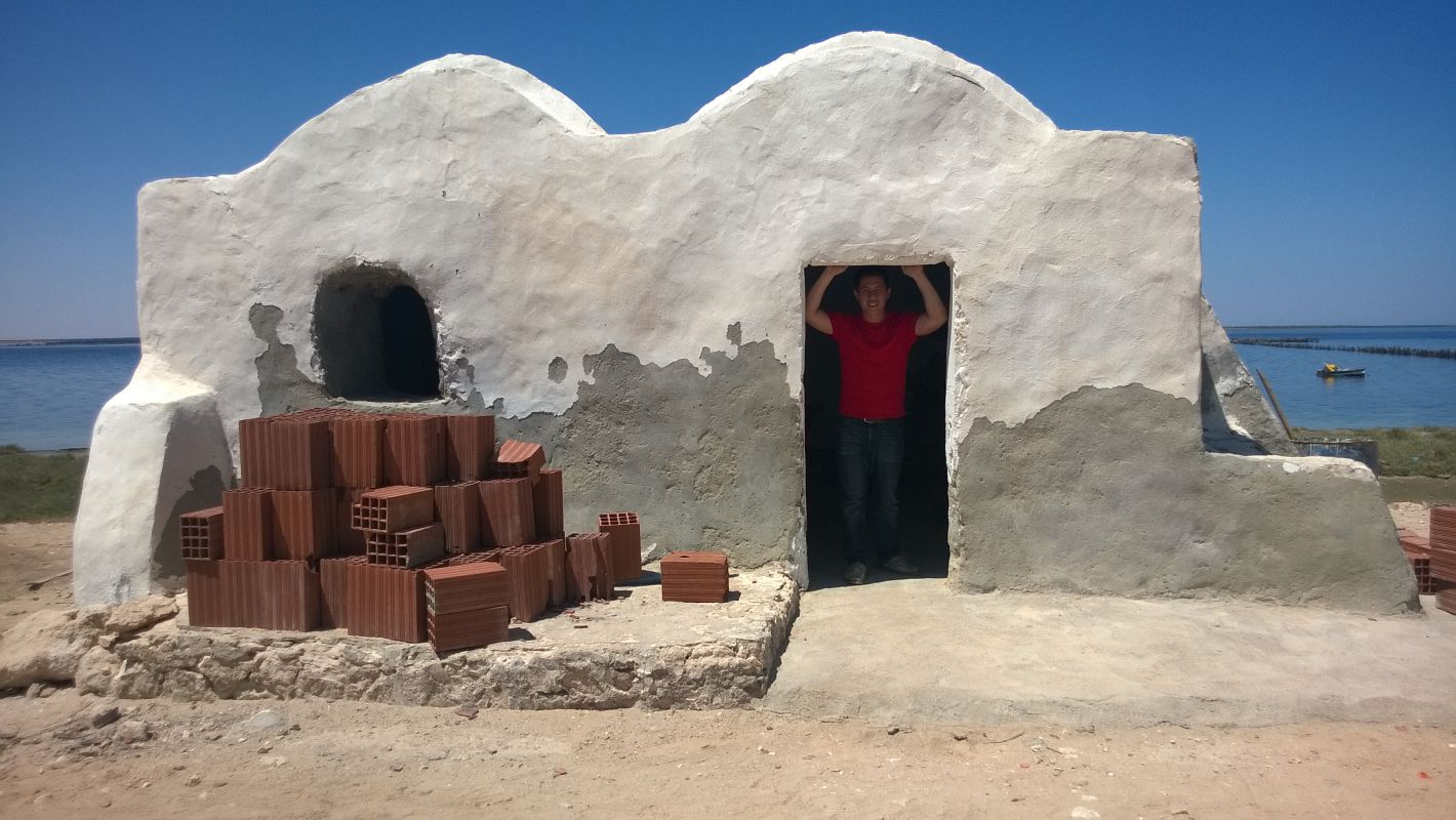 A person in a red shirt stands in the doorway of a weathered white building, an original Star Wars set, beside blue water.