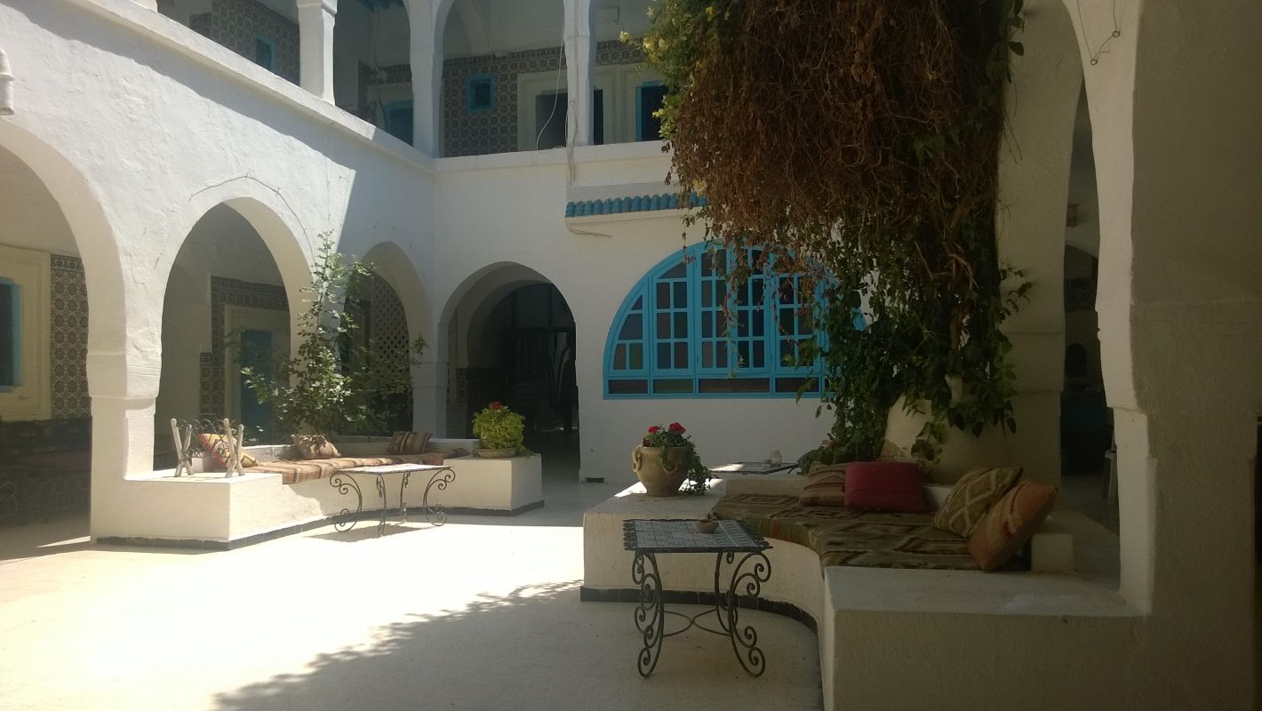 A sunlit Tunisian courtyard with white arched walls, blue windows, and cushioned seating.