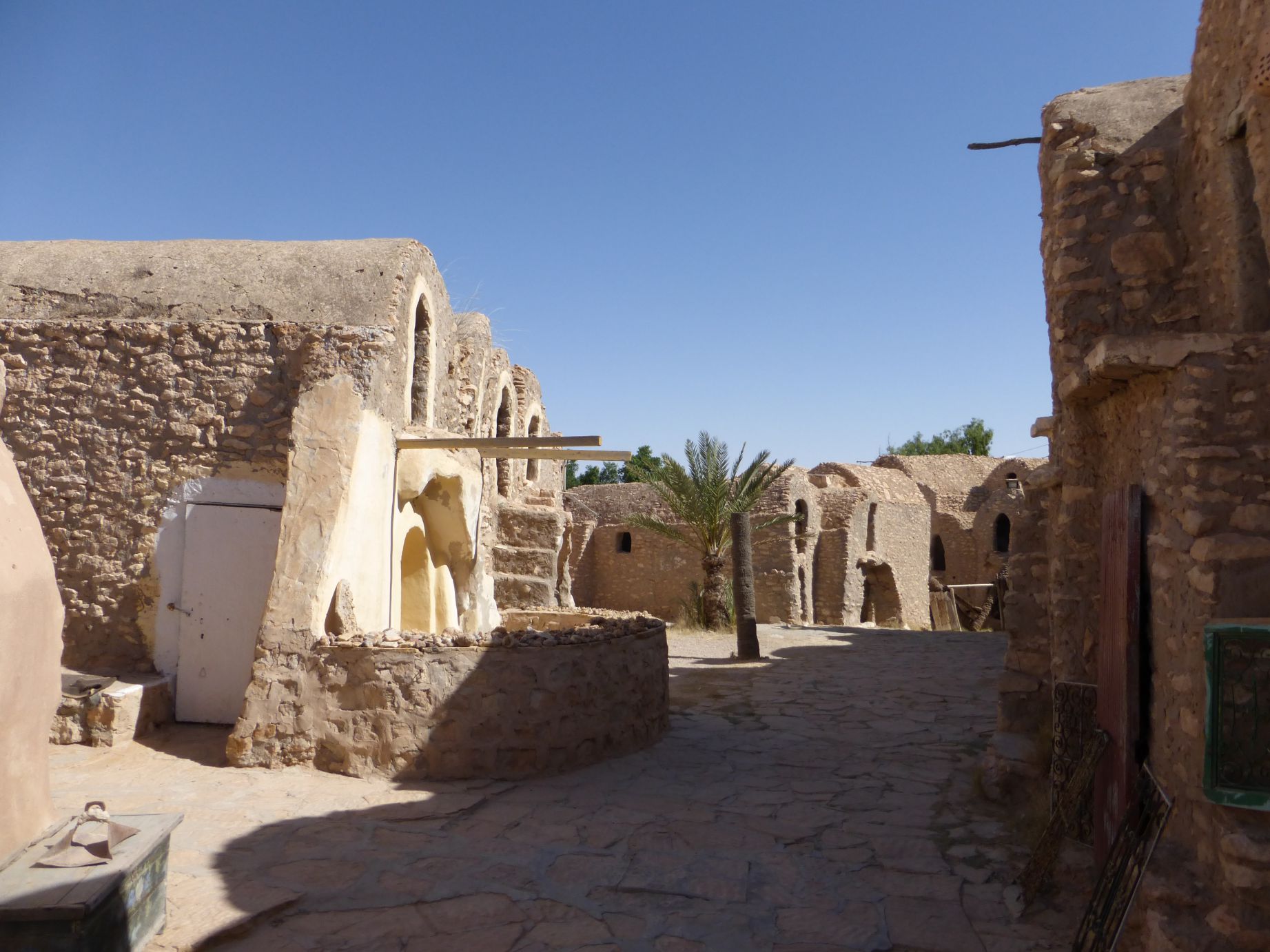Rustic stone buildings with rounded roofs and a palm tree under a clear blue sky, part of a Star Wars film set.