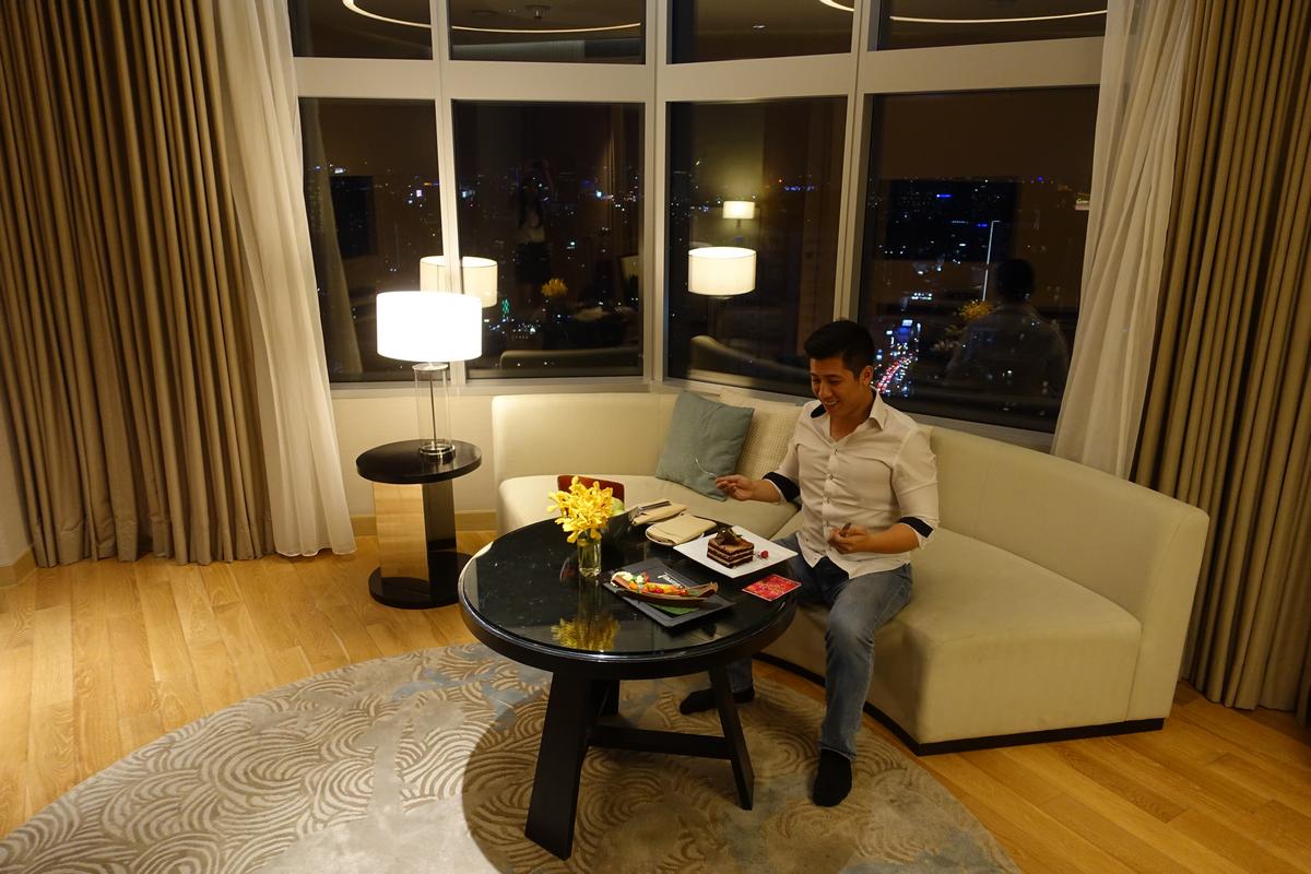 A man enjoys dessert in a modern hotel room overlooking a city at night.