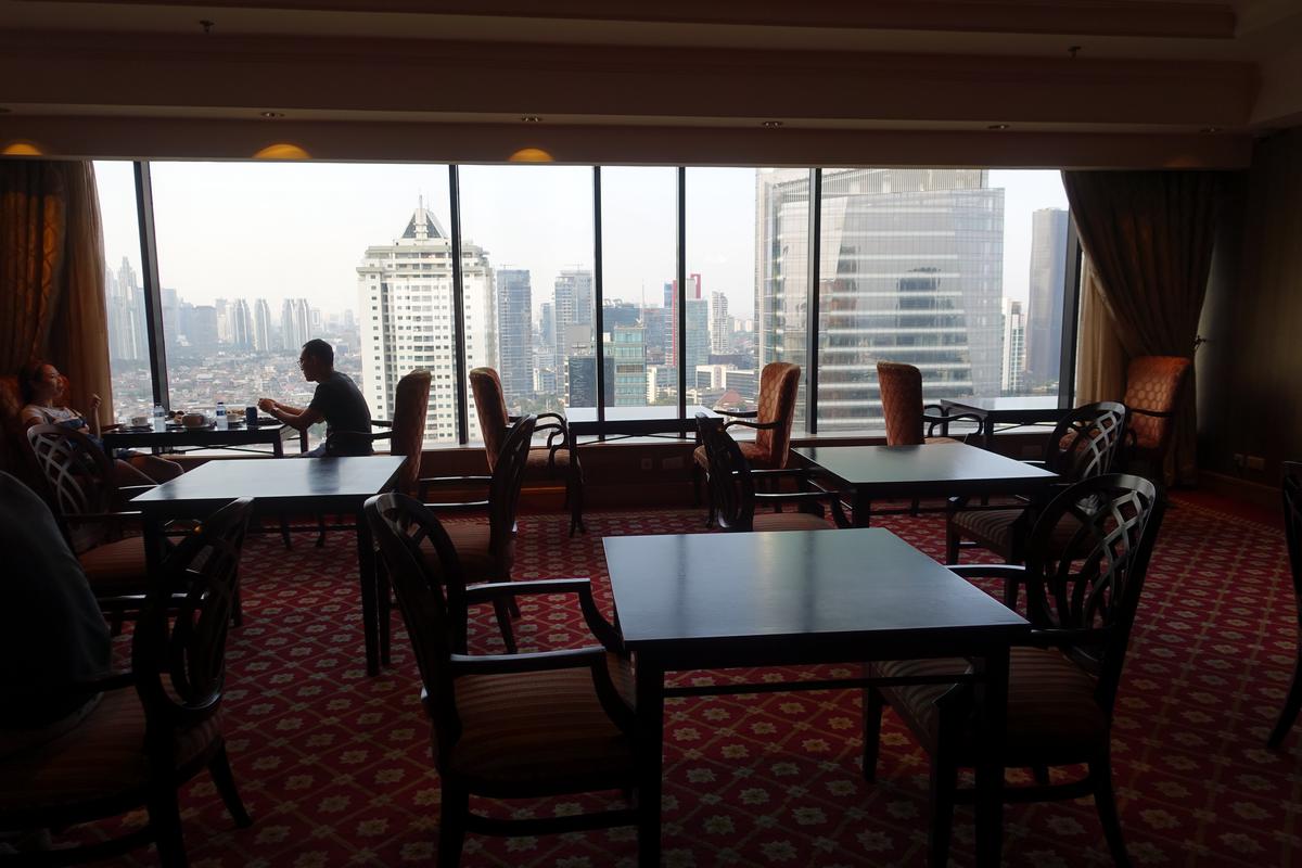 A high-rise hotel lounge offers a panoramic city view, with two guests seated by the window.