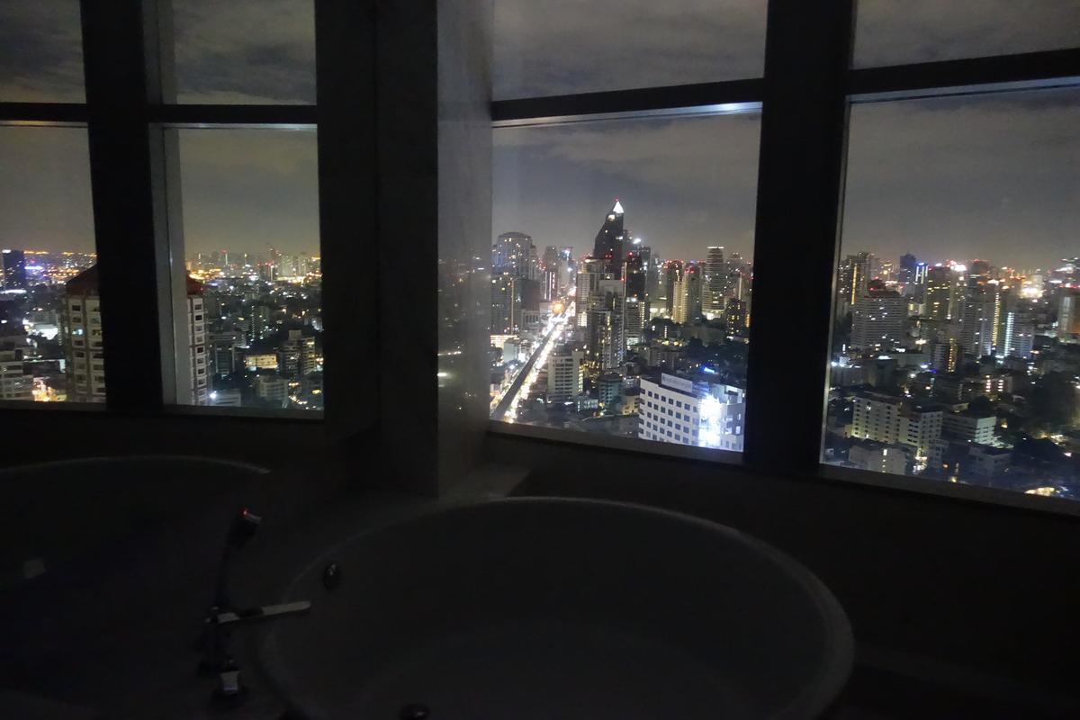 Two bathtubs in a dark room overlook a brightly lit city skyline at night through a large window.