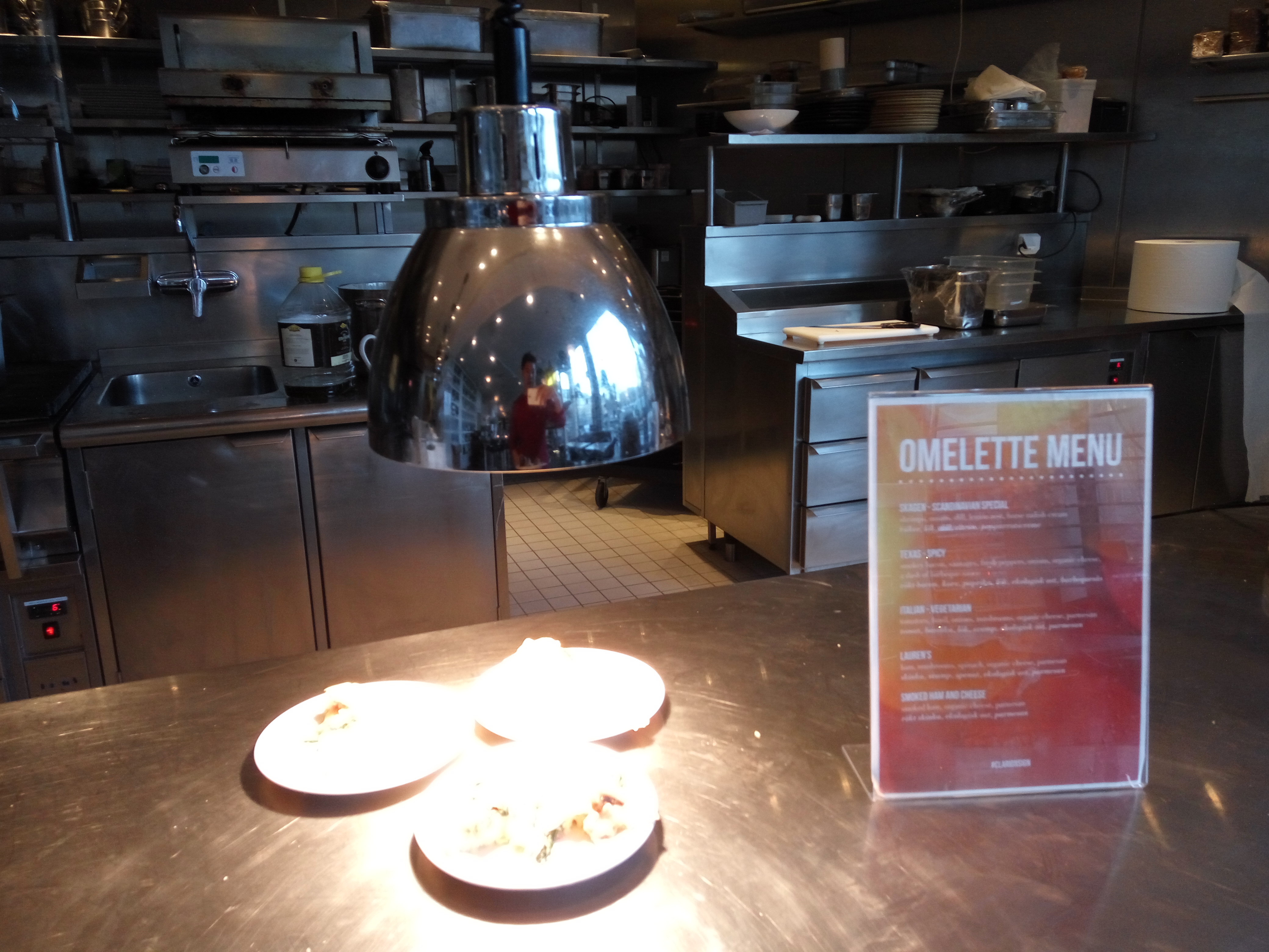 An omelette menu and plates of food sit on a stainless steel buffet counter under a heat lamp.