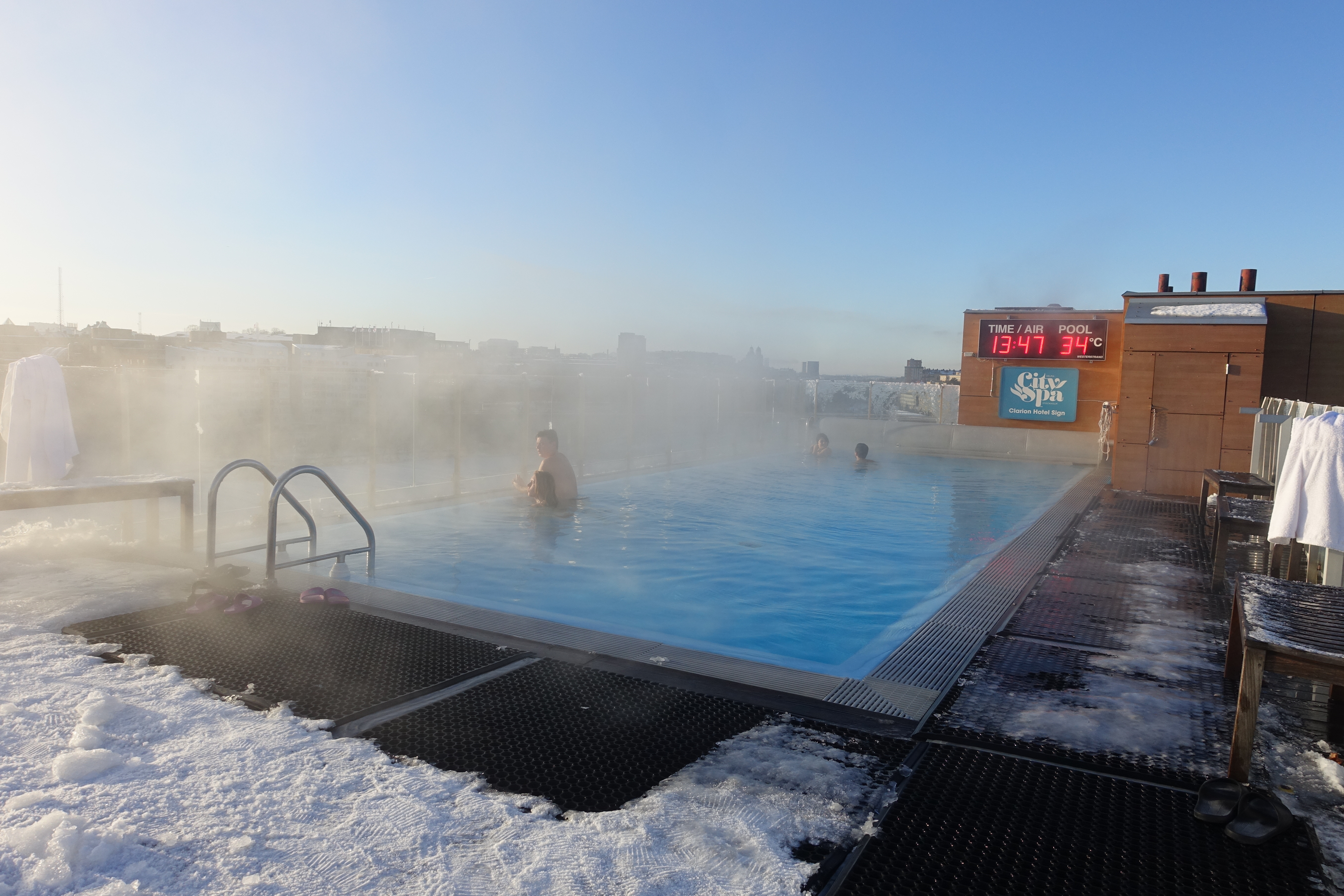 People relax in a steaming 34°C outdoor rooftop pool surrounded by snow, with a city skyline in the background.