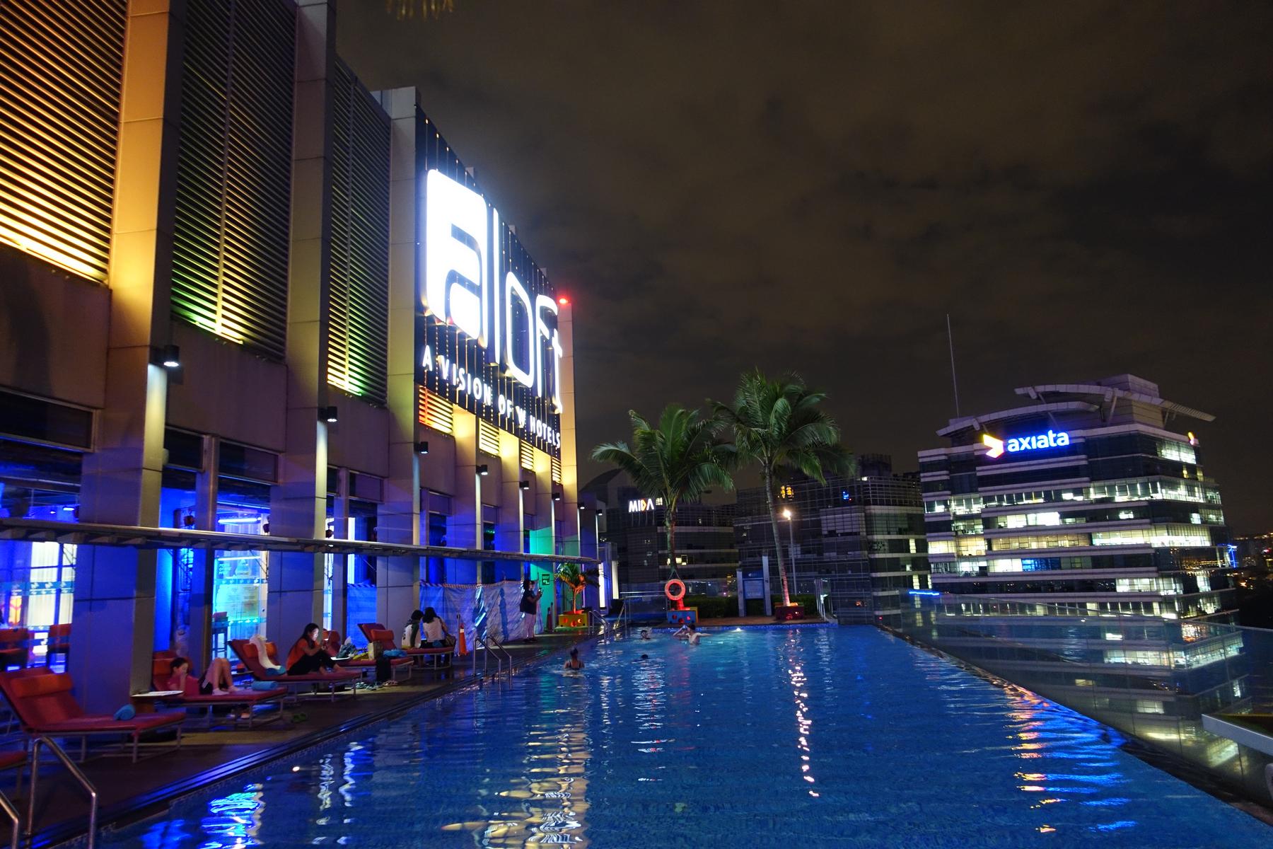 Rooftop infinity pool at the Aloft hotel at night with swimmers and illuminated city buildings.