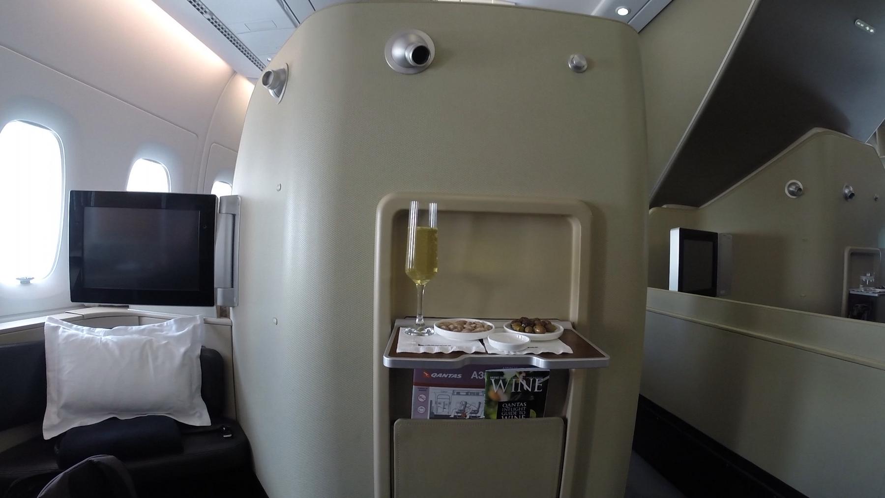 A Qantas A380 first class suite with a glass of champagne, snacks, and a personal entertainment screen.